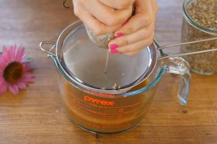Hands squeeze a cheesecloth bundle of an alcohol-based tincture over a fine mesh strainer.