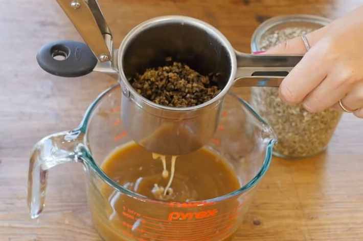 A hand holds a potato ricer full of plant matter over a glass measuring cup. This potato ricer is straining the tincture.