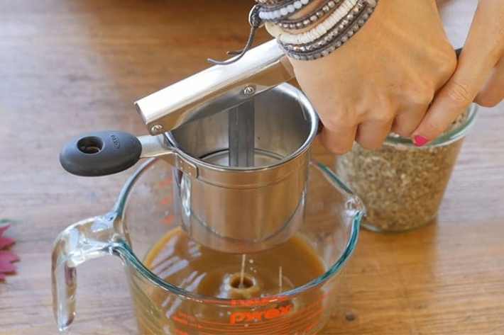 Hands use a potato ricer to strain an alcohol-based tincture of plant matter.